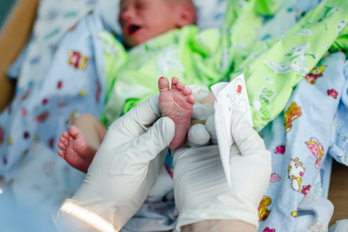 A doctor with gloves administers a newborn screening blood test on a baby in a green blanket.