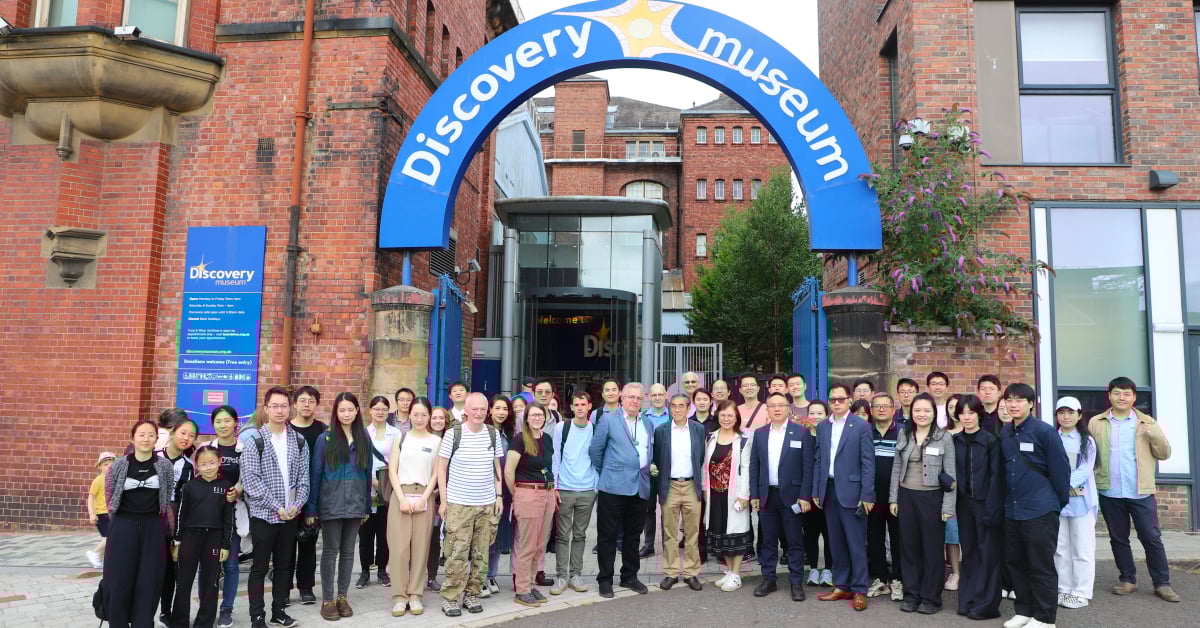 A group of symposium attendees stand before a blue arch between red brick buildings. The words 'Discovery Museum' is inscribed on the arch in white lettering.