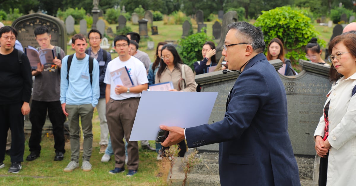 A man holding a piece of paper faces left, speaking to an attentive crowd in a graveyard.
