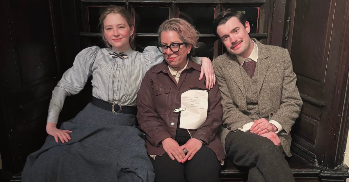 A blonde woman and brunette man in Victorian clothing sit either side of the director, a woman wearing a modern brown jacket and black glasses. The woman has her arm around the director's shoulders.
