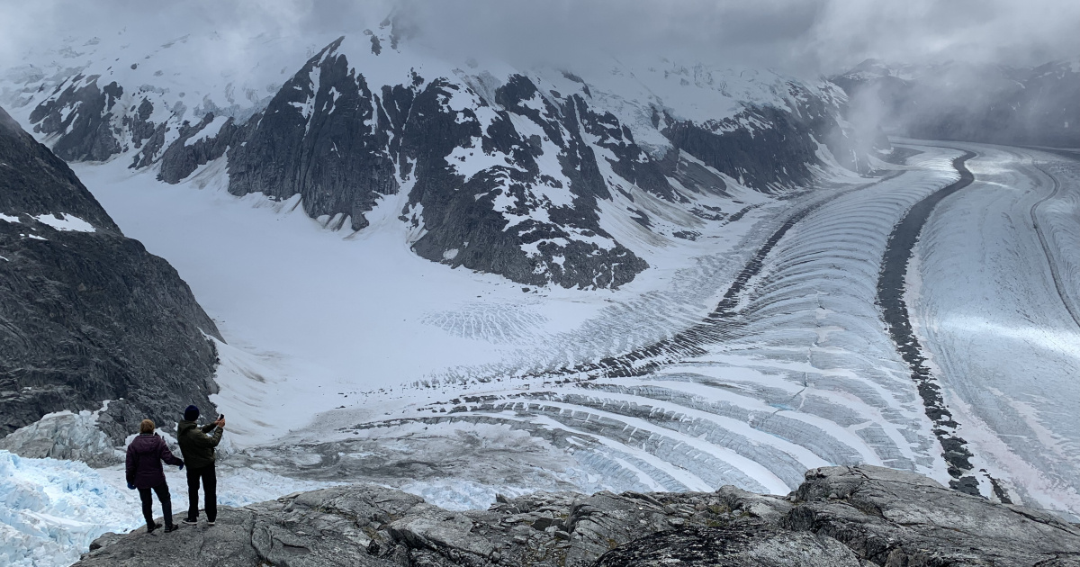 Two individuals look over the Vaughan Lewis Icefall at Gilkey Glaciar, which has created an arc-like effect of ice snaking around the snowy mountains.