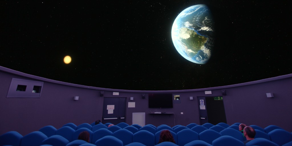 Six people sit in a dark auditorium watching a screen that shows Earth