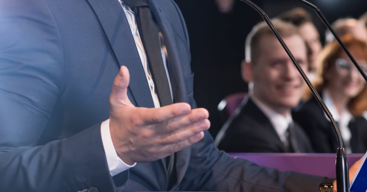 A man in a dark blue suit speaks in front of a podium while a row of people look on, smiling.