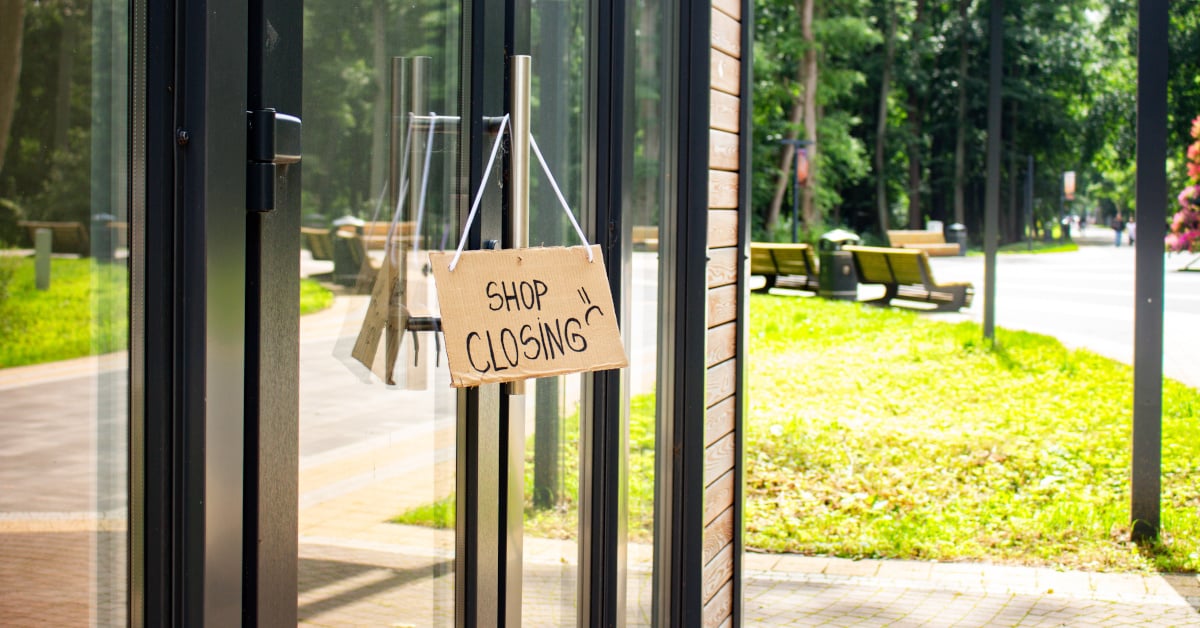 Image of a shop front with glass doors. A cardboard sign is hung on the door handle, reading 'Shop closing' with a frowning face drawn to the right of the text.