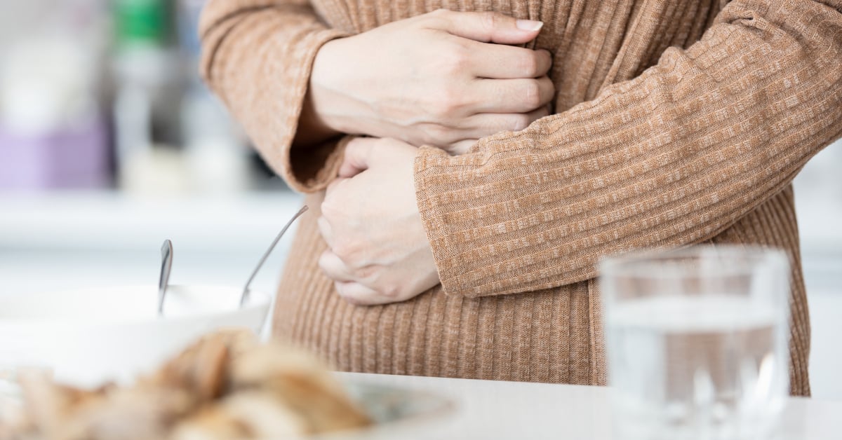 A woman in a jumper holds her stomach in pain. A variety of food is placed on the table in front of her.