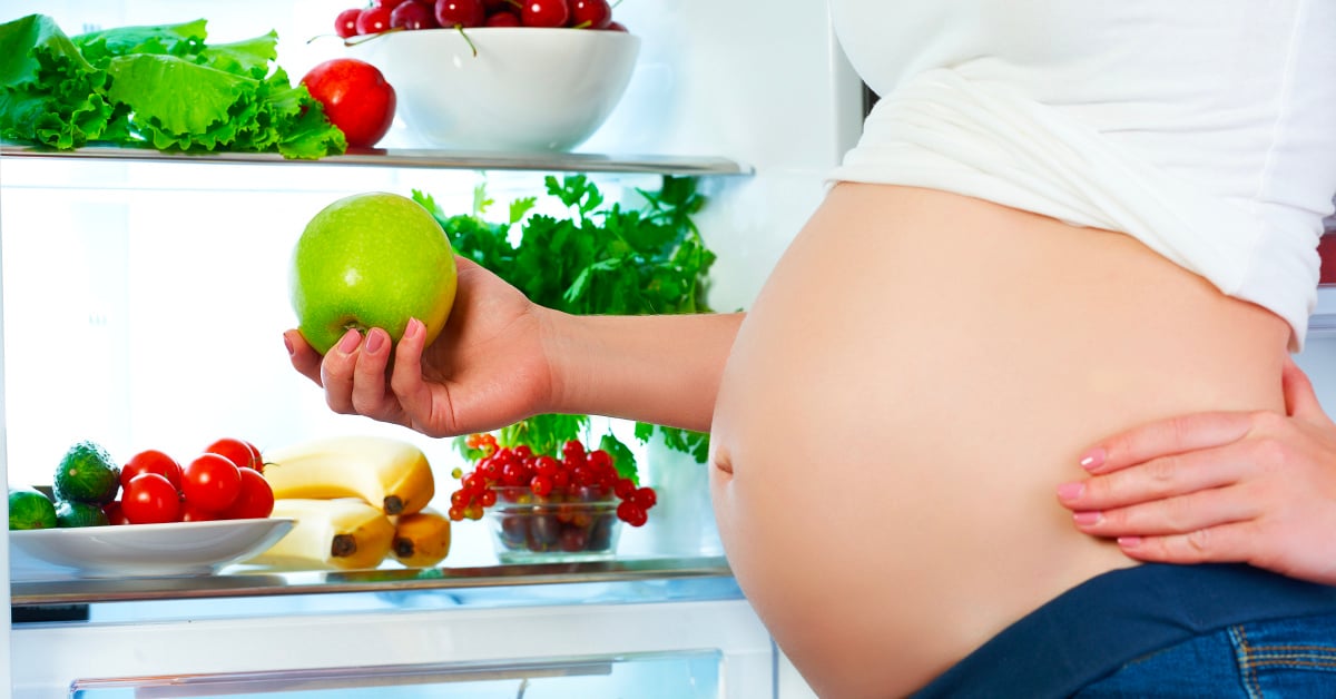 A pregnant woman facing left, standing in front of an open fridge full of groceries, holding an apple.