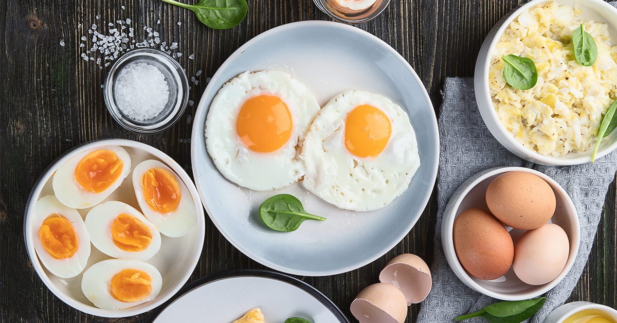 Different types of cooked eggs (poached, fried, boiled etc) on plates and in bowls on a table, as taken from above.