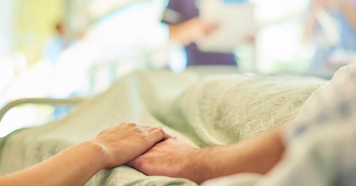 A person lying in a hospital bed holds the hand of someone sitting next to them. A nurse stands in the background, her image blurred and out of focus.