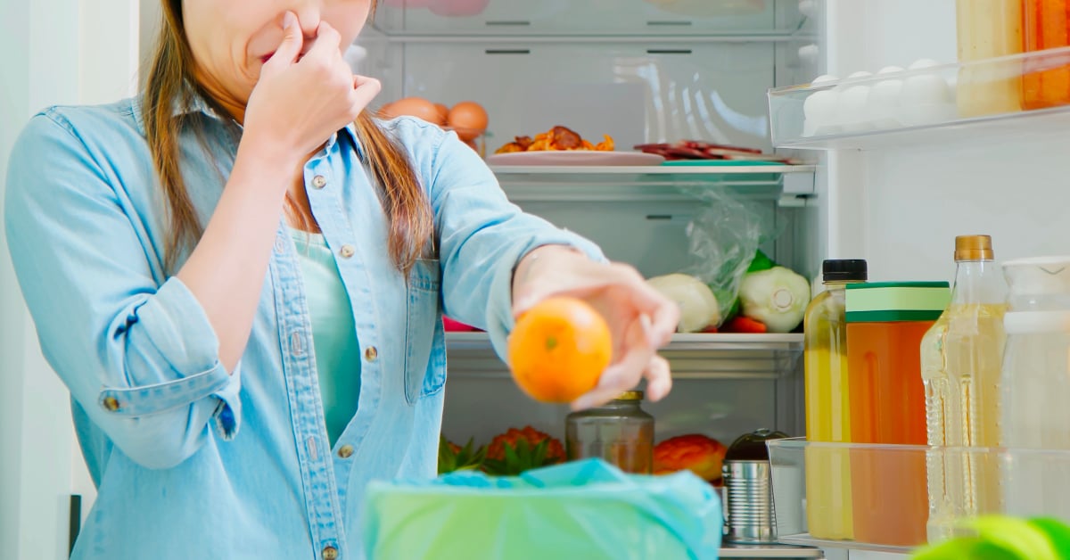 A woman in a blue shirt stands in front of an open fridge. She holds her nose while throwing an expired orange into a waste bin.
