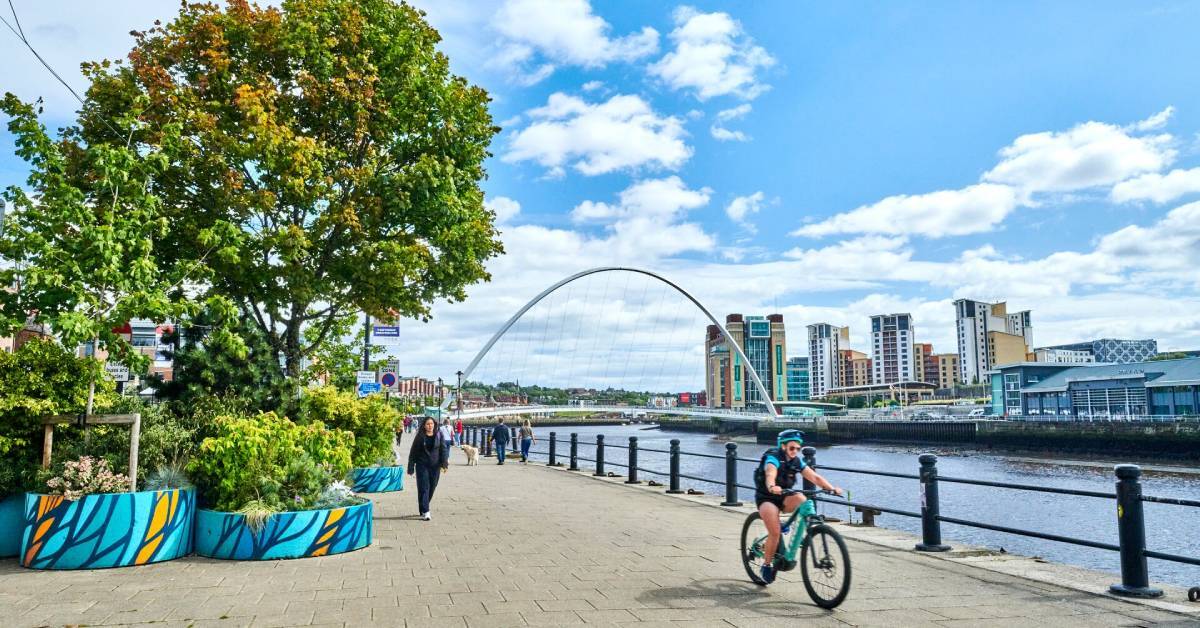 photo shows a sunny Newcastle upon Tyne, with the Millenium Bridge behind and a cyclist enjoying the quayside
