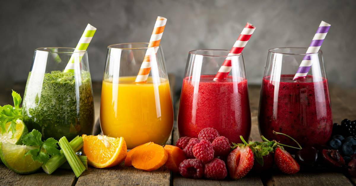 image shows four glasses of fruit juice smoothies, surrounded by a display of free fruit