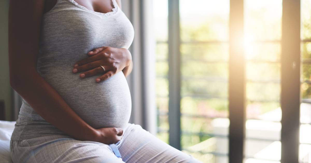 photo shows a pregnant woman holding her stomach and sitting on the edge of a bed